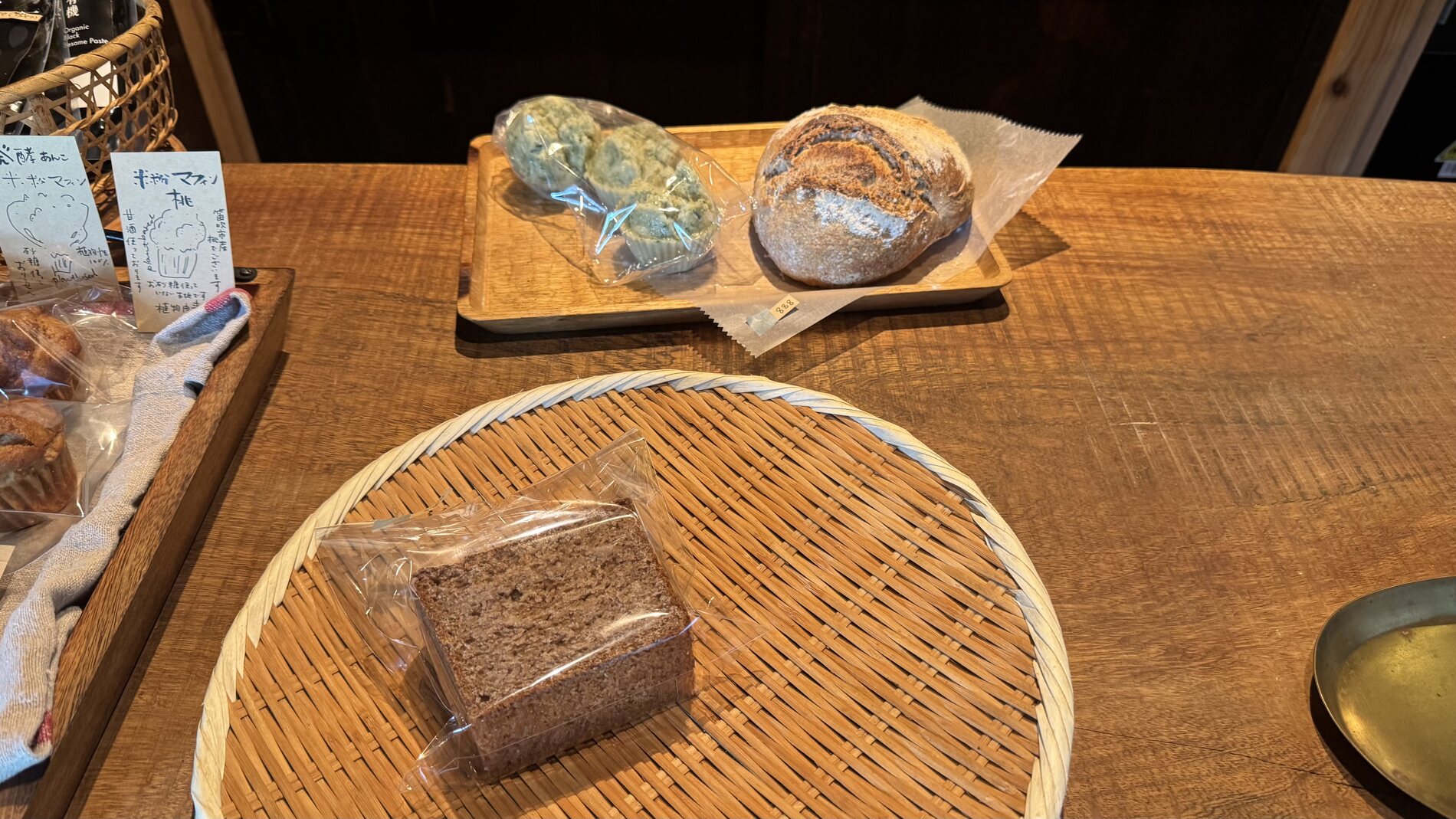 Breads bought at Pandian on Lake Kawaguchi laid on woven bamboo trays - a dark rice-flour steamed bun, a rye loaf and wrapped bread, on a wooden table.