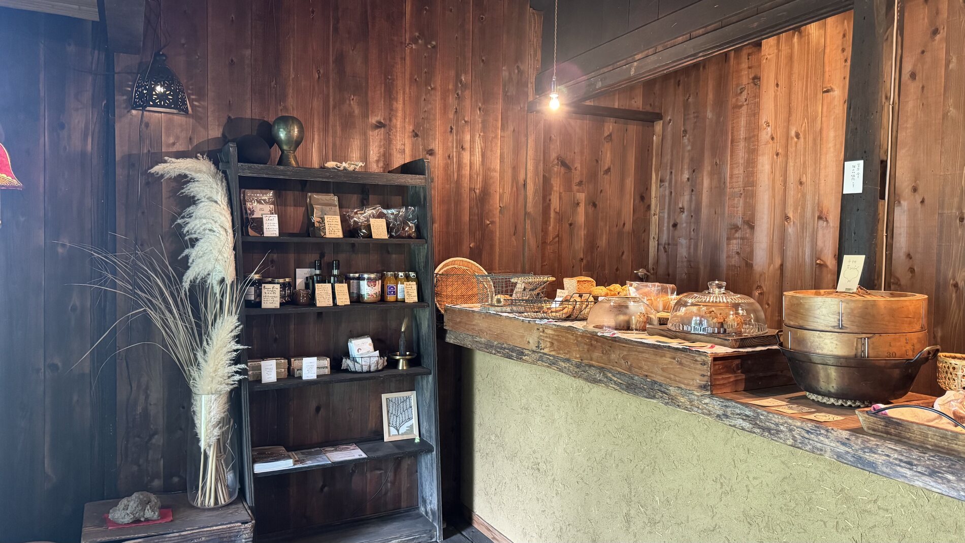 The rustic interior of Pandian bakery - a plaster-and-timber counter with dried grasses, shelves of jars and loaves of bread under warm light in Fujikawaguchiko, Yamanashi.
