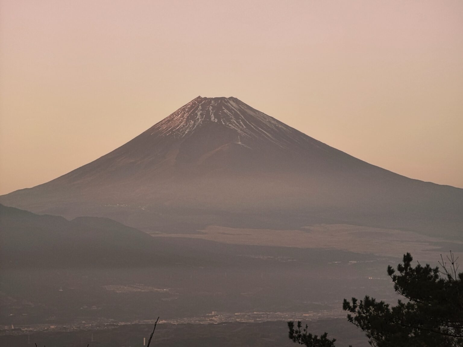 Mount Fuji in summer at dusk, its slopes nearly bare of snow under a hazy pink and orange evening sky, ridgelines and town lights faintly visible below
