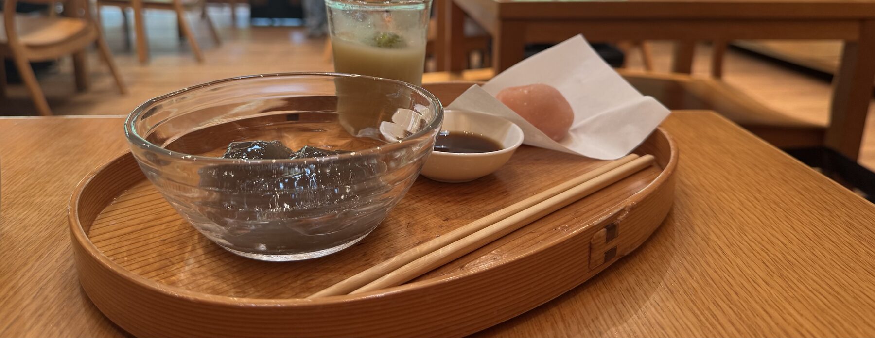 The new warabi mochi at Toraya Kobo — dark, translucent bracken-starch mochi in a glass bowl with a small dish of black-sugar syrup, served on a wooden tray in Gotemba