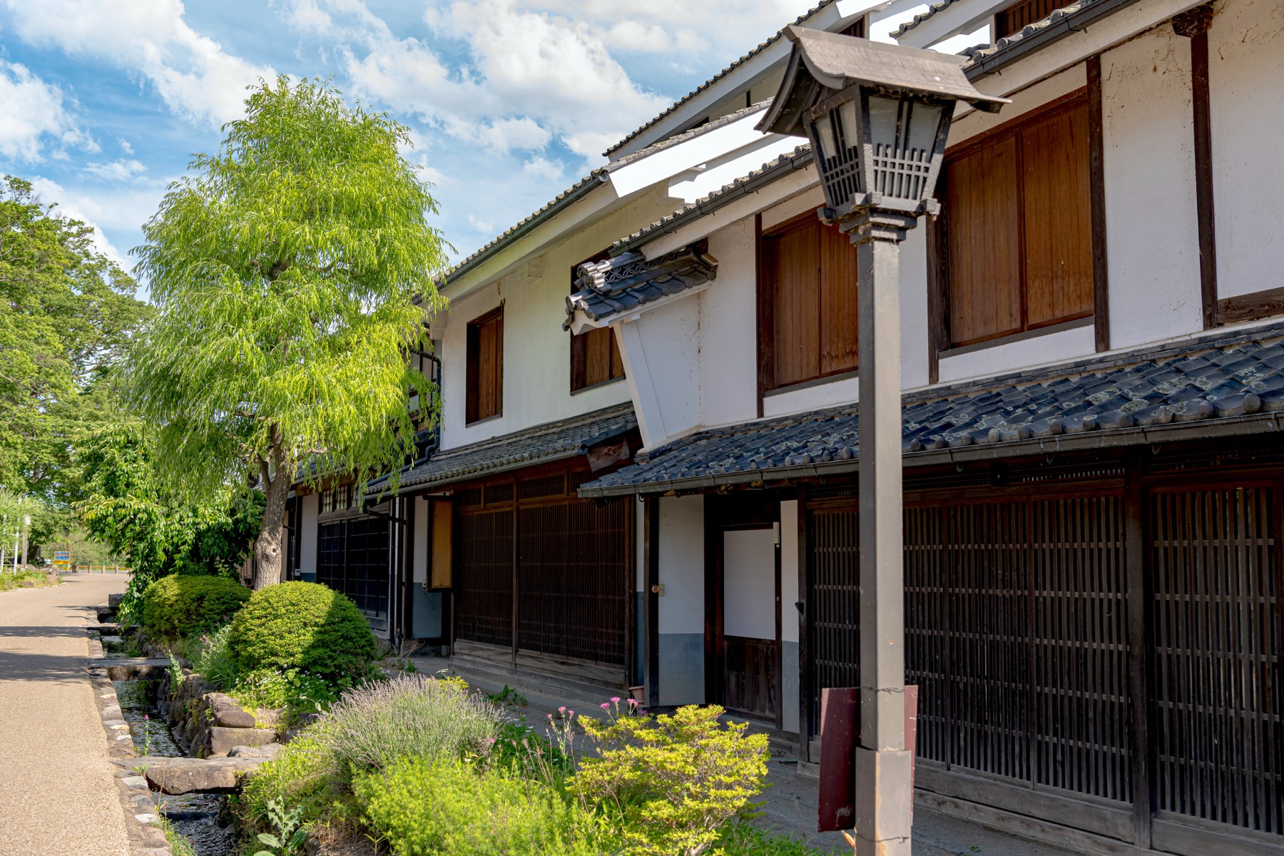 A wooden street lamp and willow beside a row of traditional lattice-fronted houses and planted flowers along the old Hokkoku Kaido street of Unno-juku Nagano