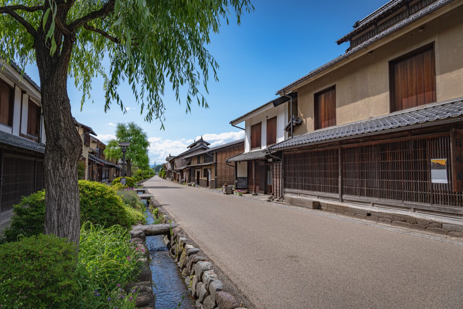 The main street of Unno-juku post town in Nagano with a stone-lined water channel and willow on the left and two-storey timber houses with white plaster and wooden lattice on the right and mountains behind