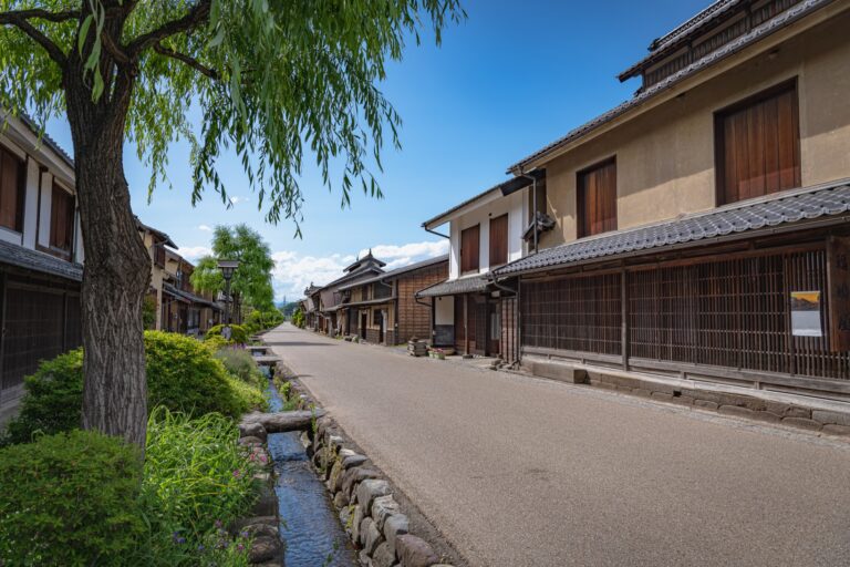 The main street of Unno-juku post town in Nagano with a stone-lined water channel and willow on the left and two-storey timber houses with white plaster and wooden lattice on the right and mountains behind