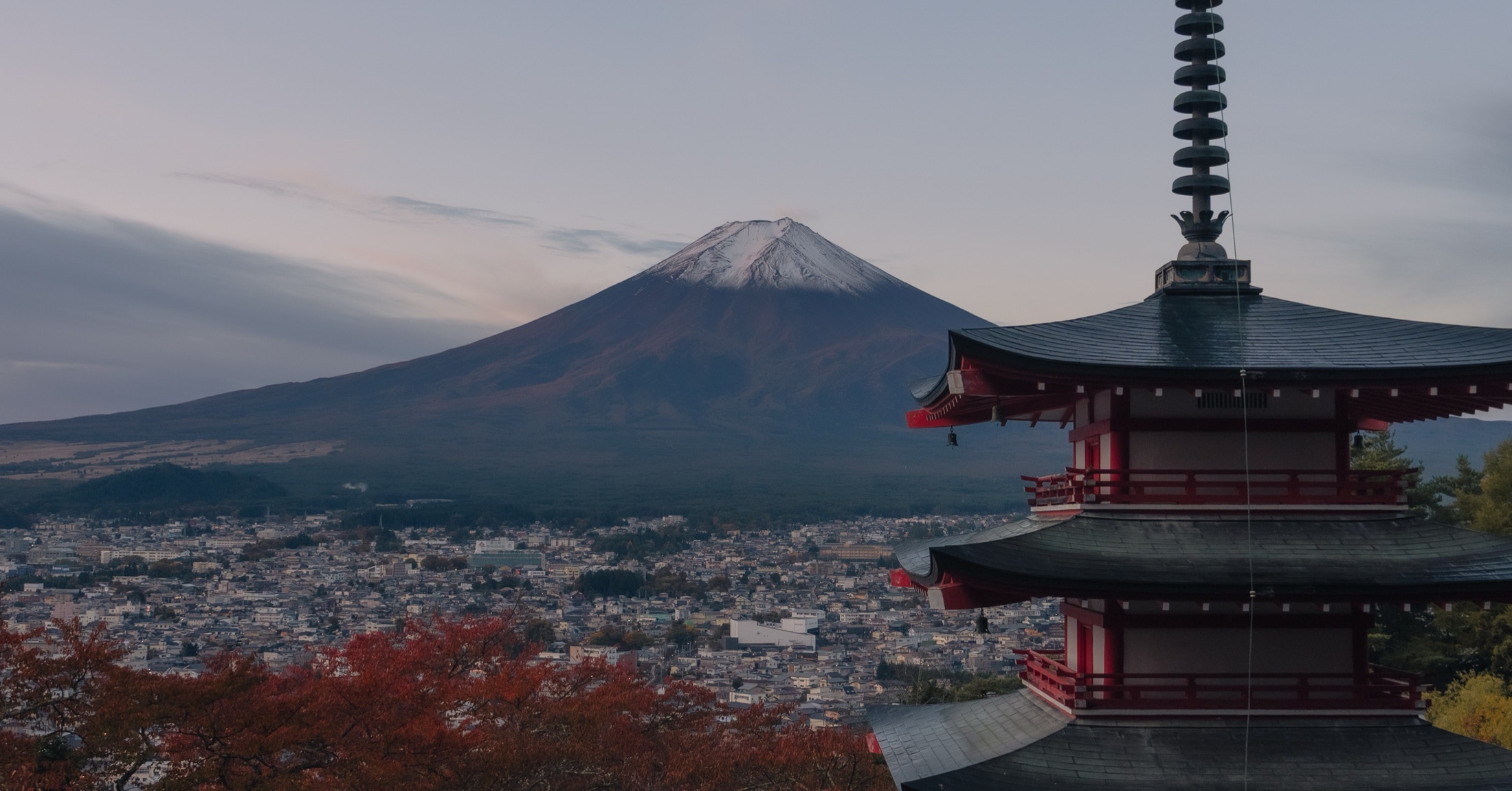 Evening view from Arakurayama Sengen Park in autumn: red and orange maple leaves in the foreground, the Chureito Pagoda on the right, and snow-dusted Mt. Fuji above Fujiyoshida city in soft dusk light