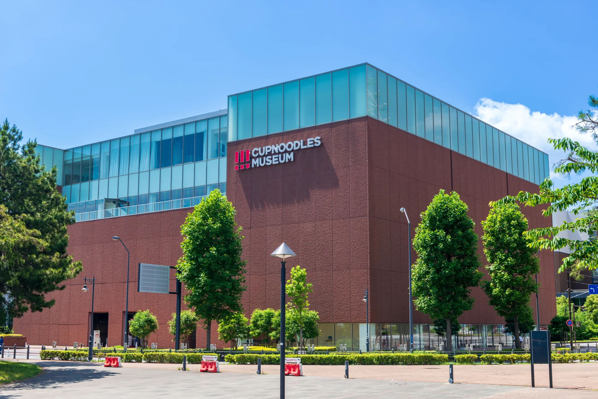 The red-brick cube exterior of the CupNoodles Museum in Yokohama Minatomirai under a clear blue sky, with the CUPNOODLES MUSEUM logo mounted on the corner of the facade
