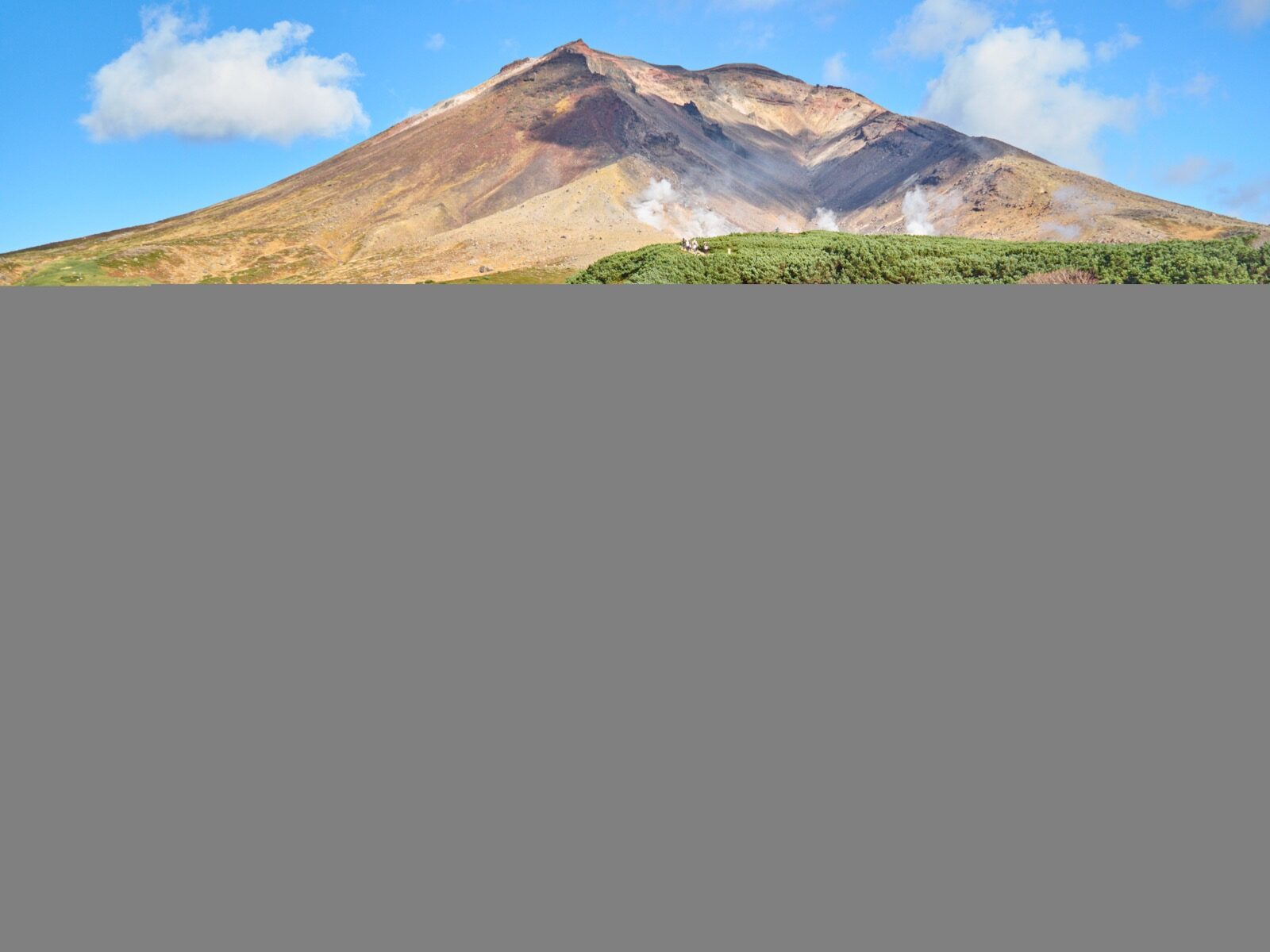 Wide alpine autumn landscape in Daisetsuzan, Hokkaido, with red and orange ground cover, dark green conifers, and rugged volcanic peaks under scattered cloud in late September.