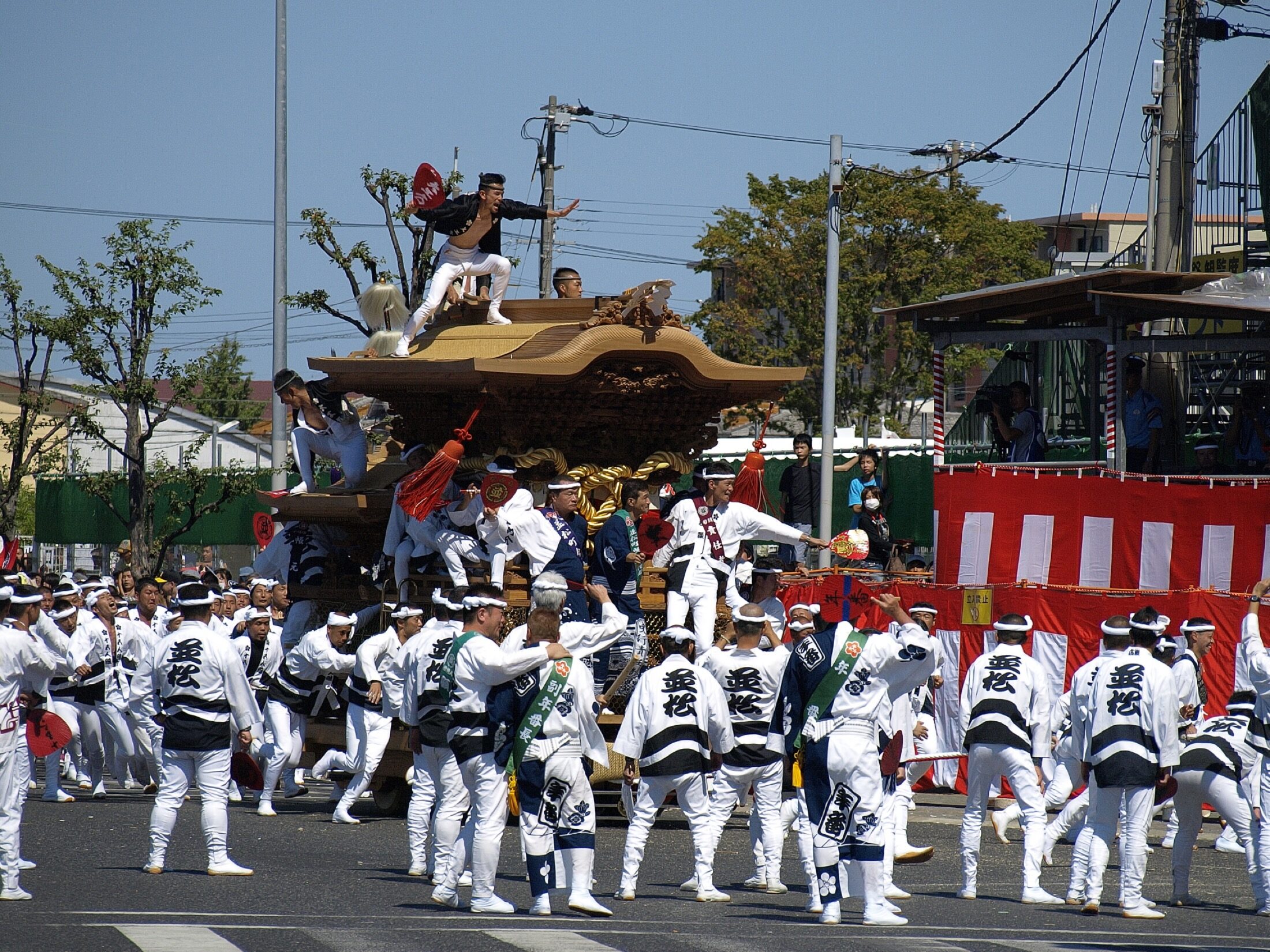 Teams in matching happi coats pulling a large wooden danjiri float at speed through the streets of Kishiwada, Osaka, by day