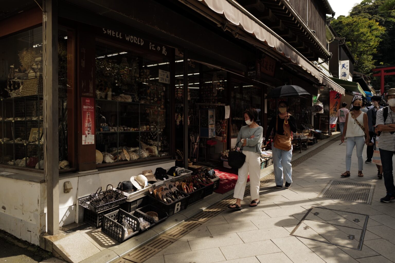 Shoppers walking up the narrow Benzaiten Nakamise shopping street on Enoshima island past a traditional shell craft shop, with the red torii gate of Enoshima Shrine visible at the top of the lane