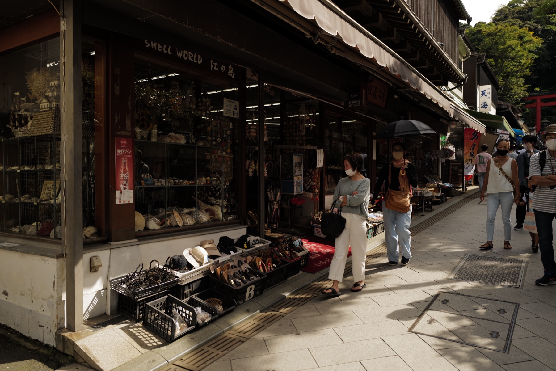 Shoppers walking up the narrow Benzaiten Nakamise shopping street on Enoshima island past a traditional shell craft shop, with the red torii gate of Enoshima Shrine visible at the top of the lane
