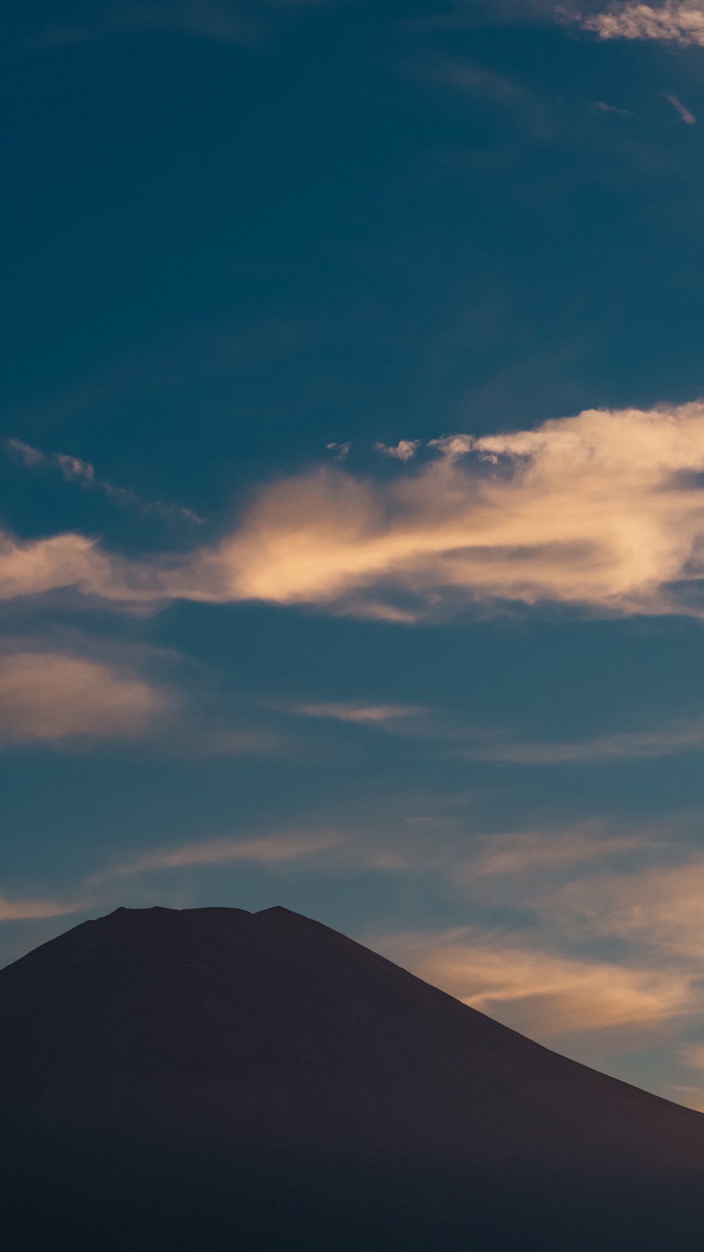 The dark silhouette of Mt Fuji fills the lower frame beneath streaks of gold and cream cloud in a deepening blue evening sky, seen from the Hana no Miyako area near Lake Yamanaka.