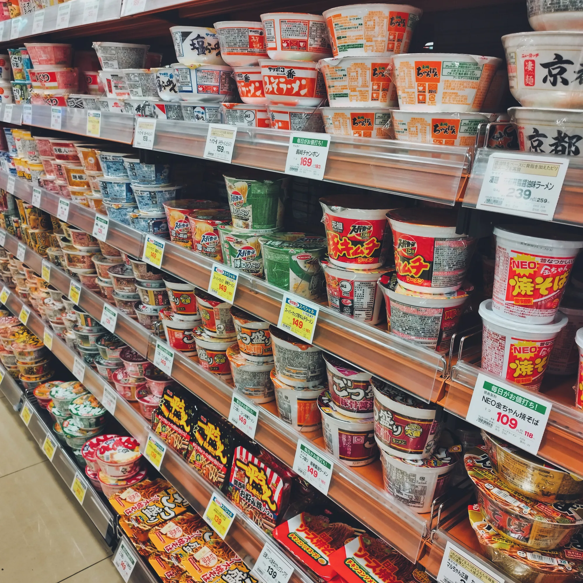 A Japanese supermarket aisle stacked floor to ceiling with dozens of different cup noodle brands and flavors, each shelf edge marked with yellow price tags well under 200 yen