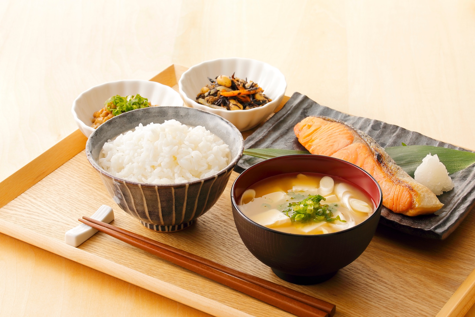 A traditional Japanese breakfast set on a wooden tray with grilled salmon, rice, miso soup with tofu, simmered hijiki seaweed, and a small bowl of natto with green onions — the kind of spread served at ryokan and hotel buffets