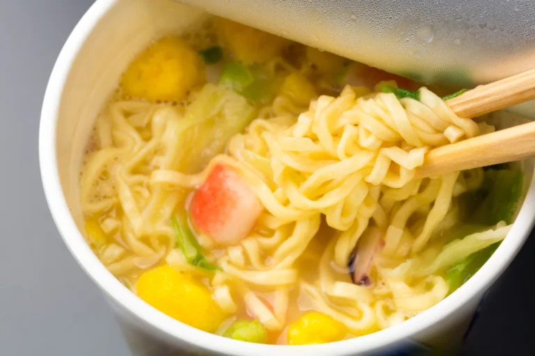 Chopsticks lifting yellow ramen noodles out of a steaming Japanese cup noodle, with kamaboko fish cake, egg and green vegetables floating in the hot soy-based broth