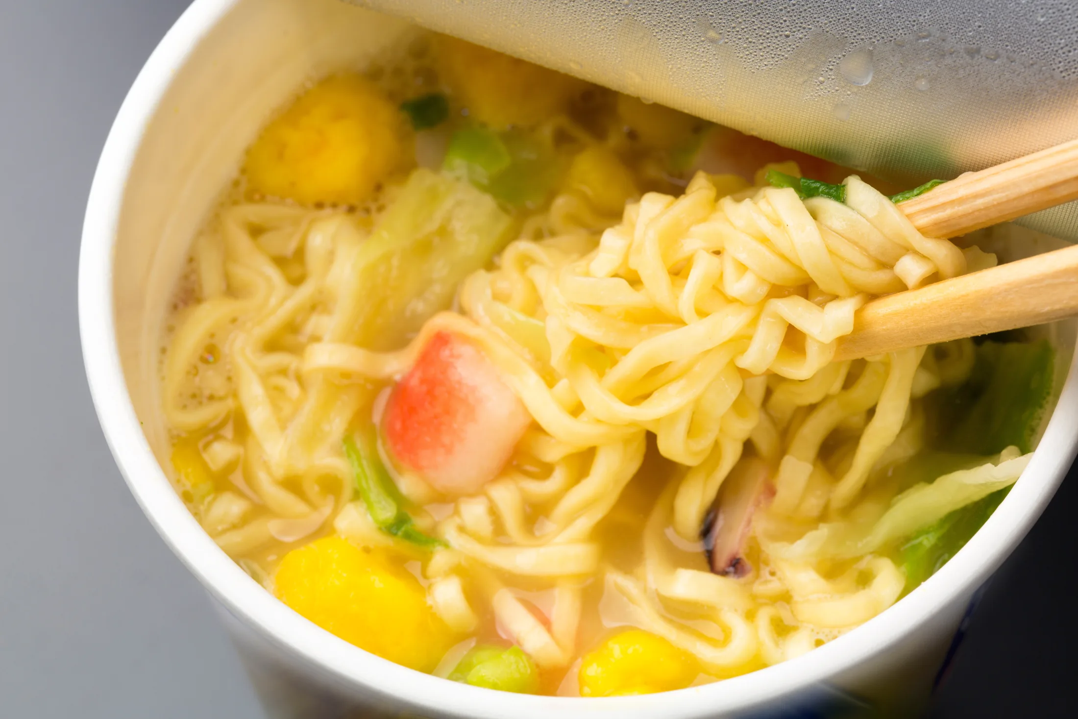 Chopsticks lifting yellow ramen noodles out of a steaming Japanese cup noodle, with kamaboko fish cake, egg and green vegetables floating in the hot soy-based broth