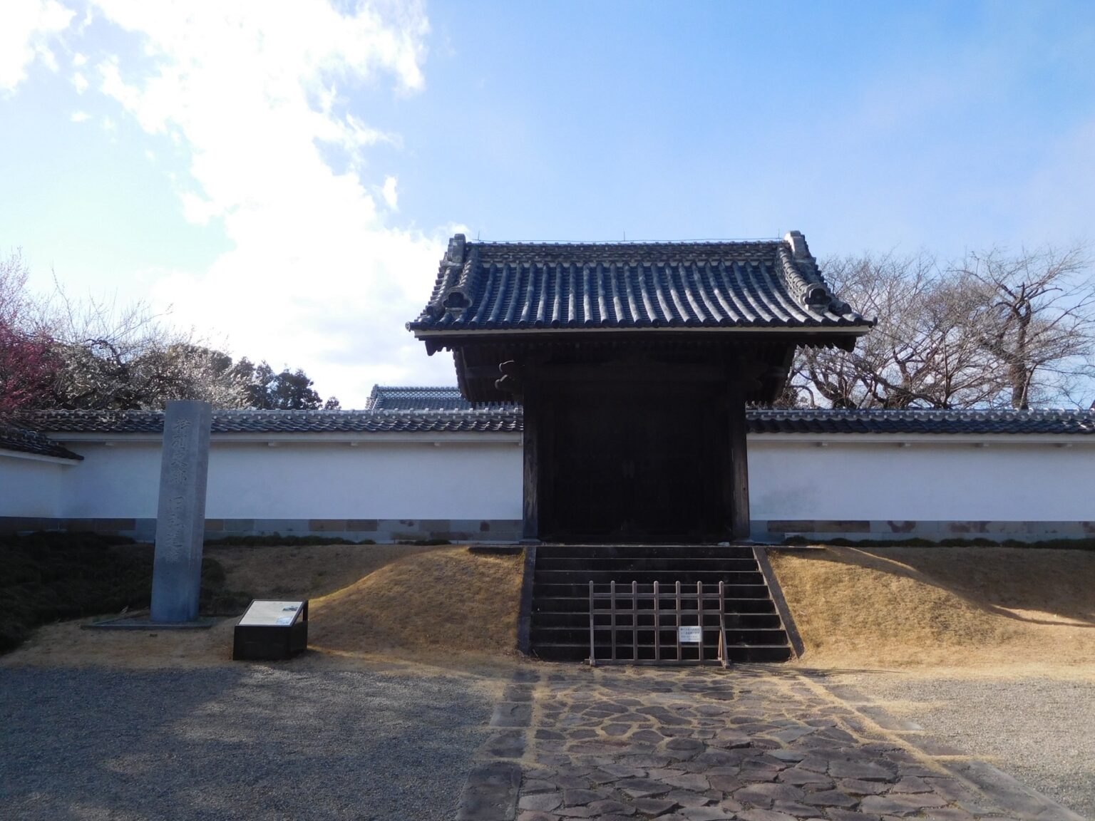 The old wooden gate of Kodokan in Mito, a dark tiled-roof samurai school gate from 1841 framed by white earthen walls, with plum trees blooming pink and white behind the wall on a clear day