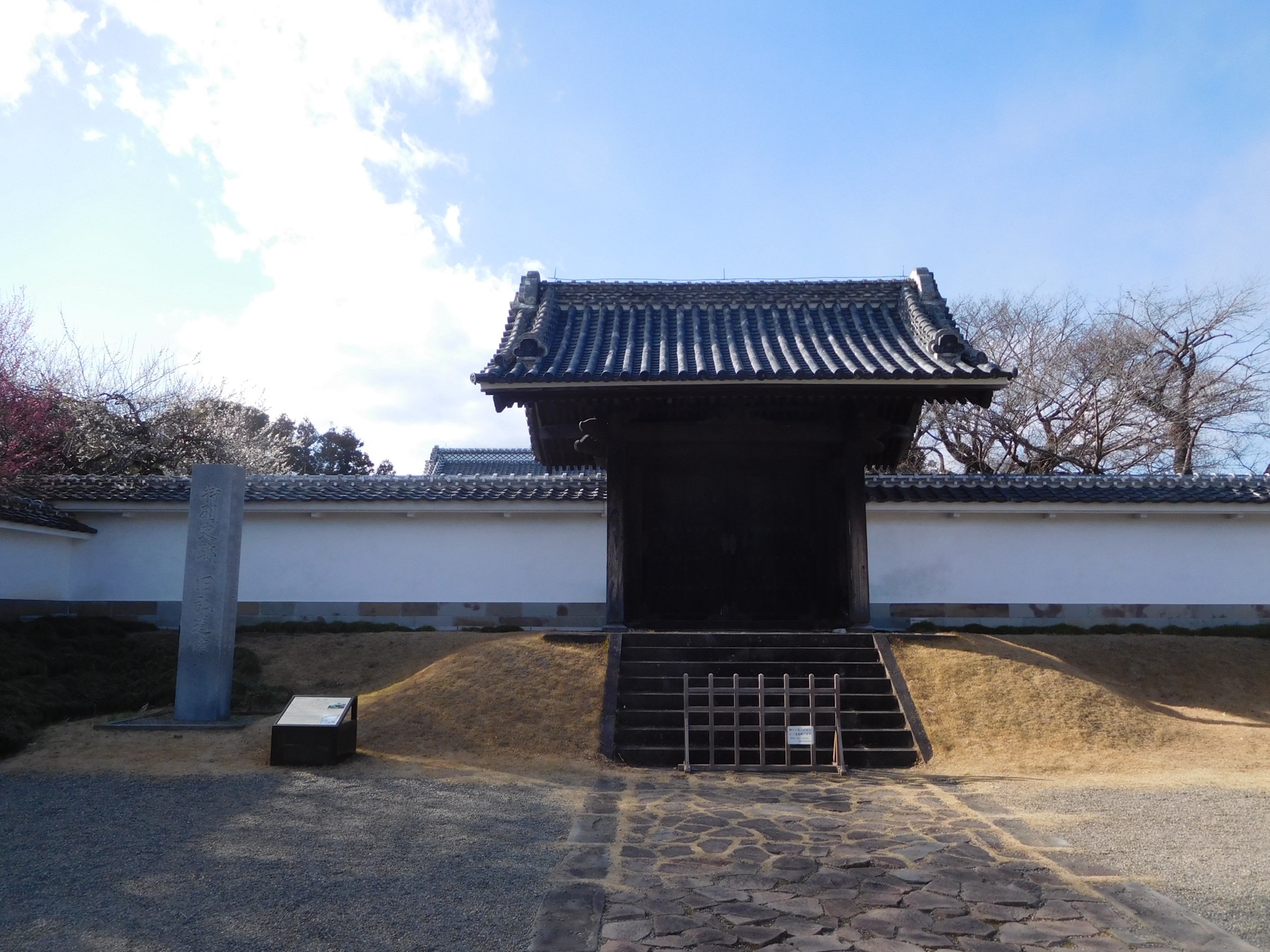 The old wooden gate of Kodokan in Mito, a dark tiled-roof samurai school gate from 1841 framed by white earthen walls, with plum trees blooming pink and white behind the wall on a clear day