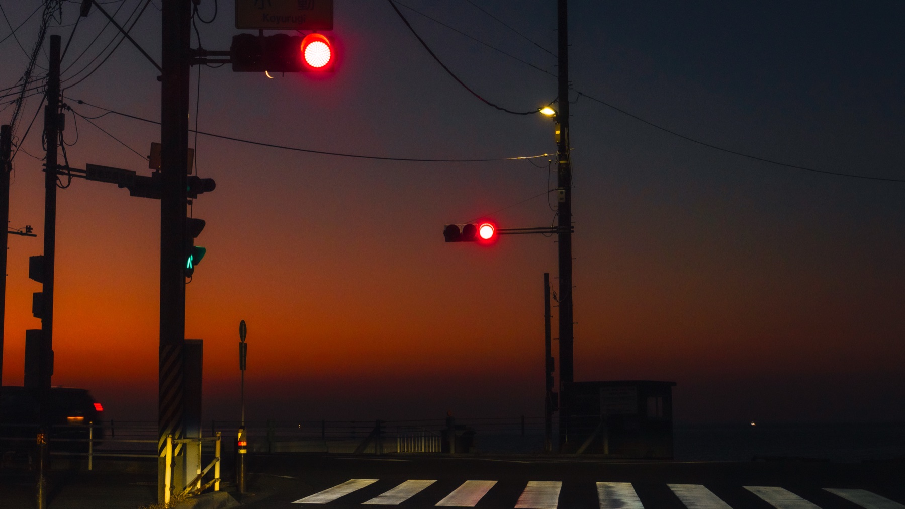 The Koyurugi pedestrian crossing on Route 134 near Shichirigahama before sunrise, red traffic signals glowing against a deep orange dawn sky over Sagami Bay with the crosswalk stripes catching the first light