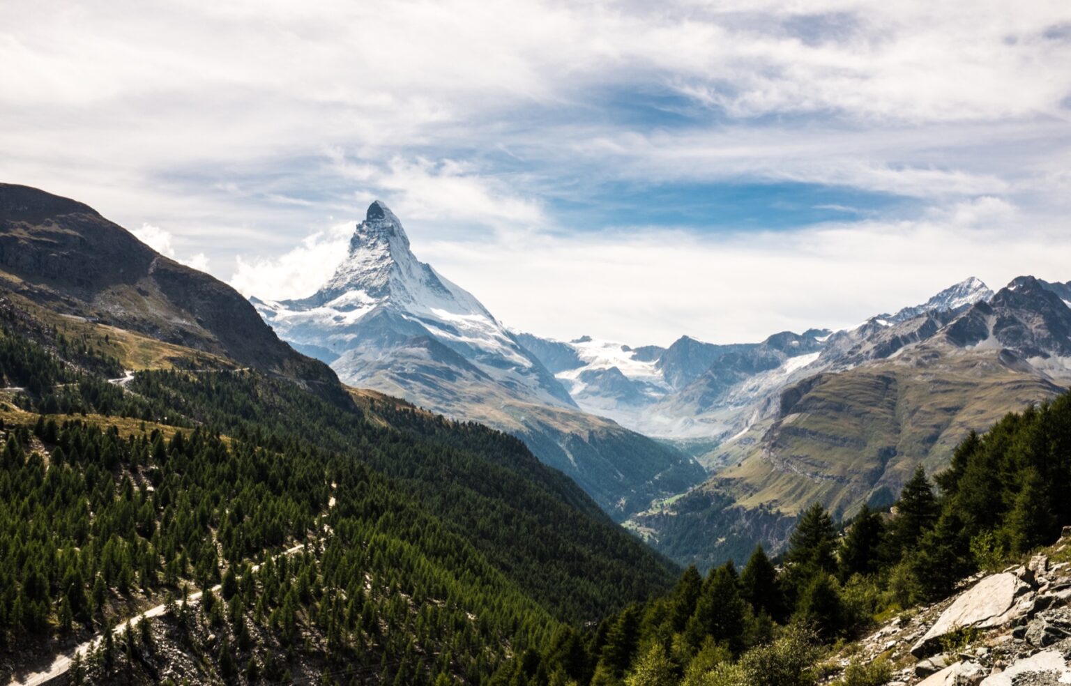 The sharp rock pyramid of the Matterhorn rising above a green alpine valley of pine forest and pasture near Zermatt in the Swiss Valais, with cloud drifting around the summit ridge.
