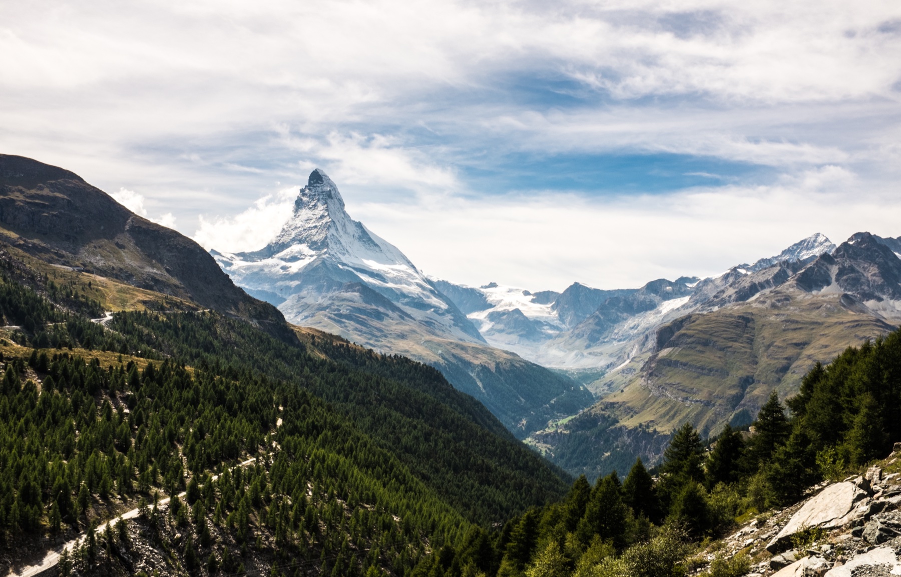 The sharp rock pyramid of the Matterhorn rising above a green alpine valley of pine forest and pasture near Zermatt in the Swiss Valais, with cloud drifting around the summit ridge.