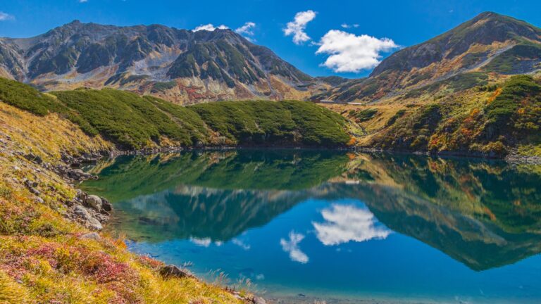 The Mikurigaike pond on the Murodo plateau of the Tateyama Kurobe Alpine Route in autumn, with red and gold alpine slopes and the Tateyama peaks reflected in still blue water at 2,450 metres