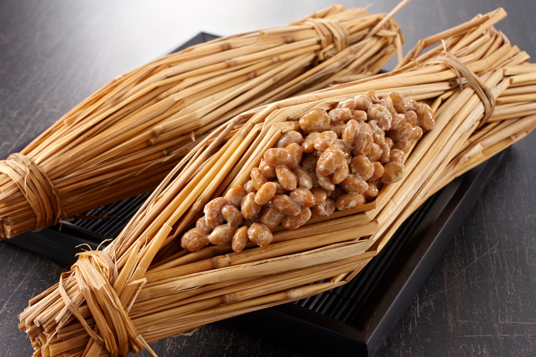 Two straw-wrapped warazuto natto bundles tied with cord resting on a black lacquer tray, the traditional Mito presentation with fermented soybeans visible through the opened straw casing