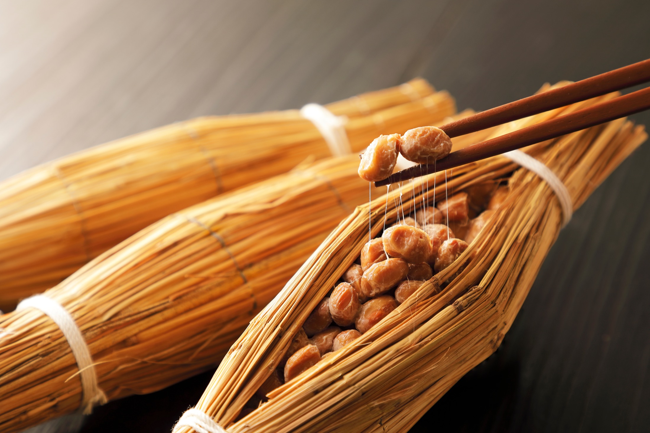 Chopsticks lifting sticky fermented natto soybeans with stretching threads from a traditional straw warazuto wrapper on a dark table, the classic Mito Ibaraki presentation of straw-wrapped natto