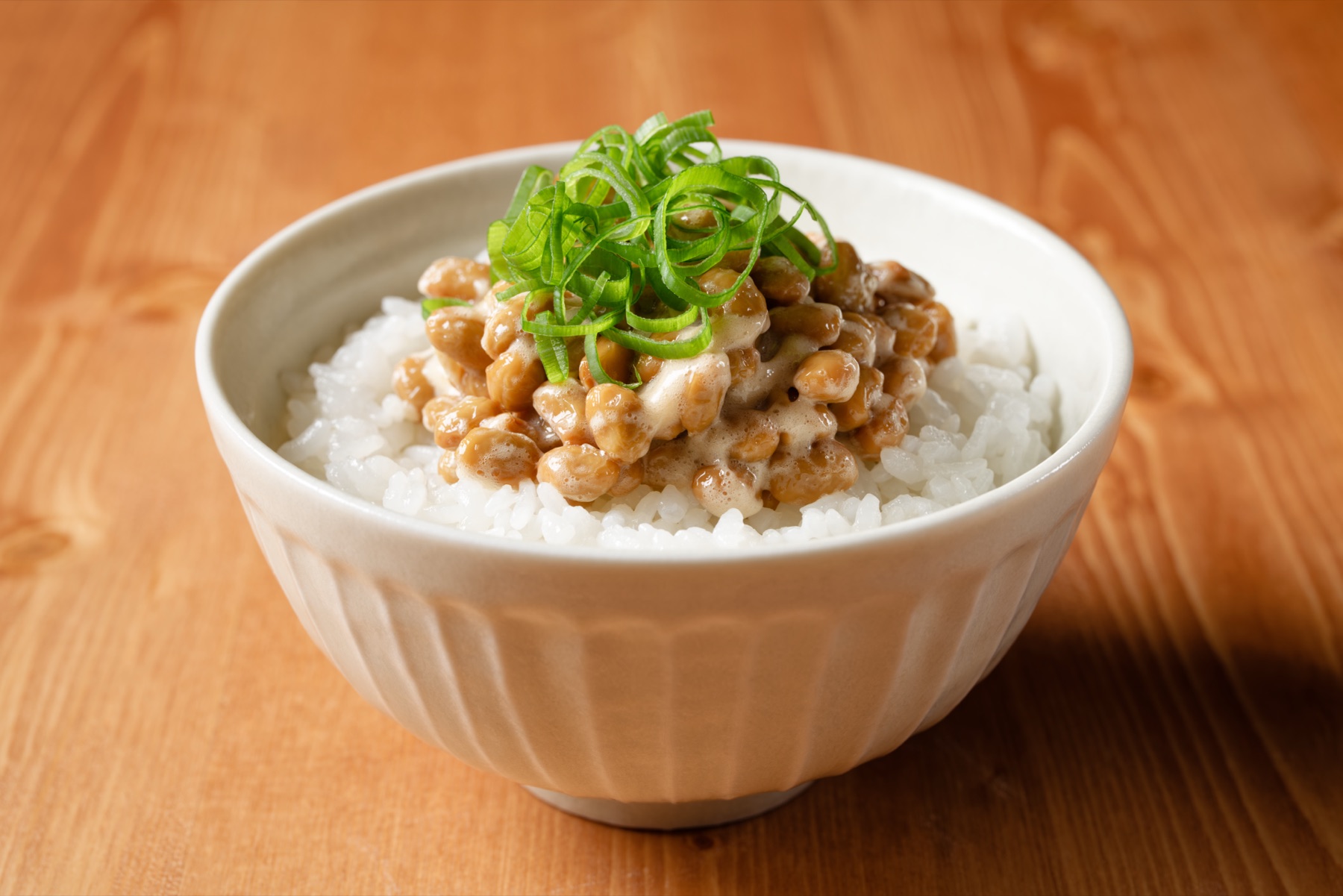 A bowl of natto gohan — fermented soybeans with sliced green onions served over freshly cooked white rice in a white ceramic bowl on a wooden table, the standard way natto is eaten for breakfast across Japan
