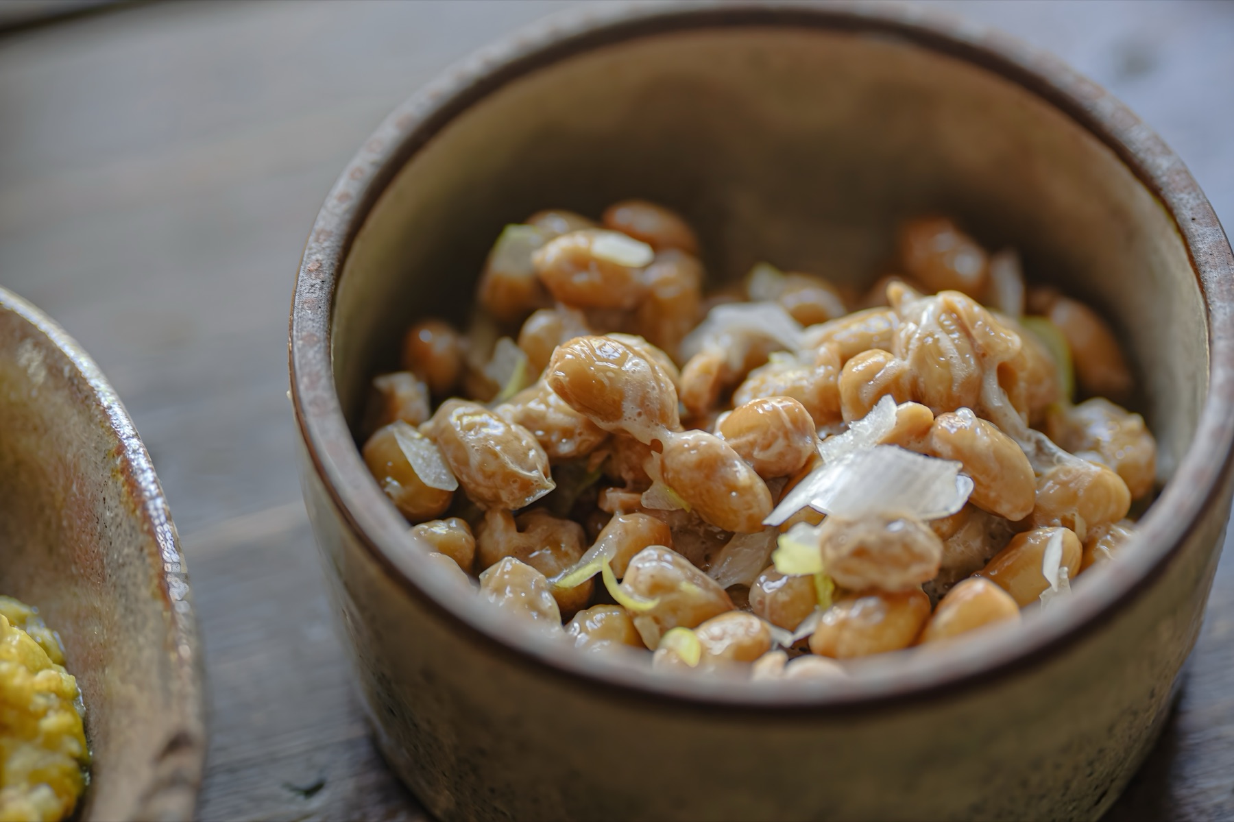 Close-up of natto fermented soybeans mixed with chopped green onions in a rustic ceramic bowl, showing the glossy coating and fine sticky threads that develop after stirring the beans thoroughly