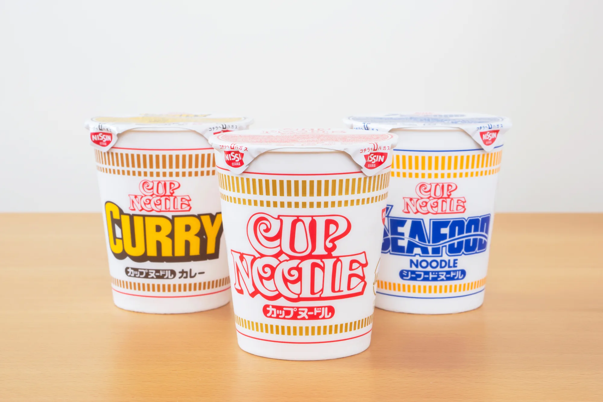 Three cups of Nissin Cup Noodle lined up on a wooden counter in Japan: curry flavor in a gold cup, the original soy-sauce flavor in the white-and-red cup, and seafood flavor in blue
