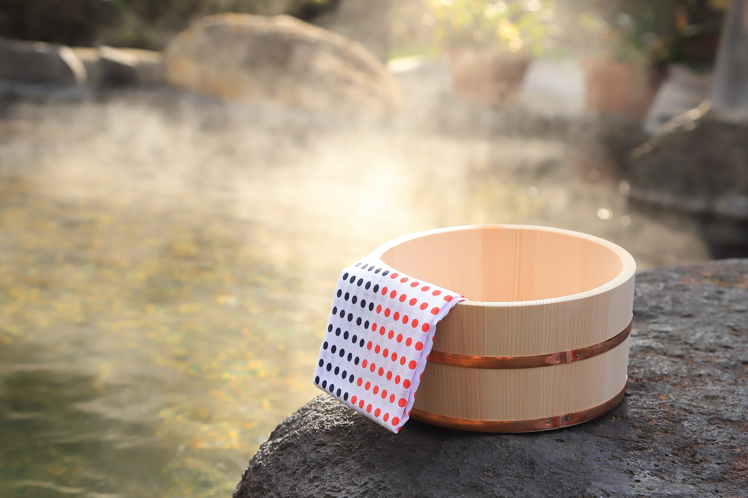 A wooden bath bucket with a folded small towel resting on a rock beside a steaming outdoor onsen rotenburo