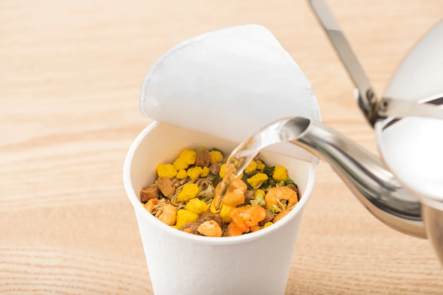 Hot water being poured from a stainless kettle into an open Japanese cup noodle showing dried shrimp, corn, egg and vegetable flakes before the three-minute wait