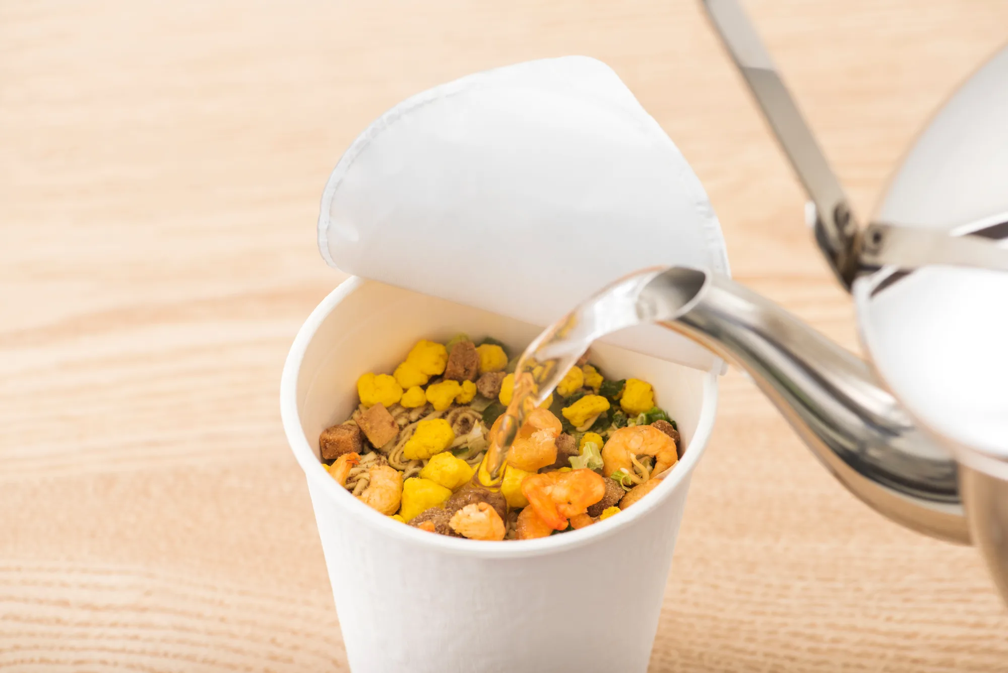 Hot water being poured from a stainless kettle into an open Japanese cup noodle showing dried shrimp, corn, egg and vegetable flakes before the three-minute wait