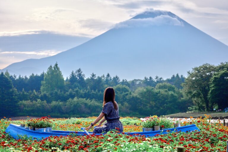 A visitor sits in a sky-blue rowboat photo spot floating in a sea of red, orange and yellow zinnias at the Rainbow Flower Festival, with Mt Fuji rising through morning haze behind the treeline at Fuji Motosuko Resort, Yamanashi.