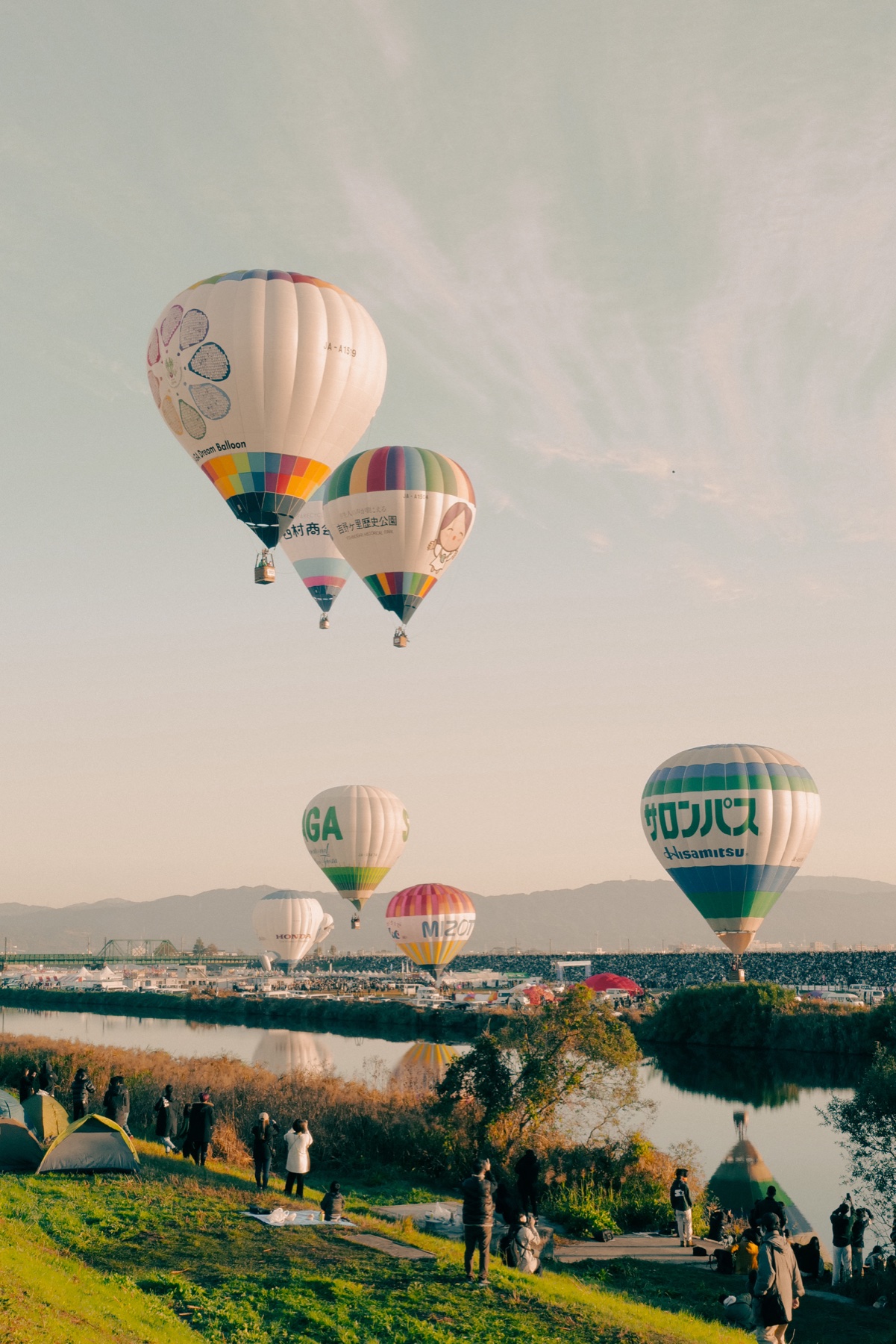 Several hot air balloons hang in the pale morning sky above the Kase River at the Saga International Balloon Fiesta, their reflections on the water, with tents and a few spectators dotted along the quiet west bank below.