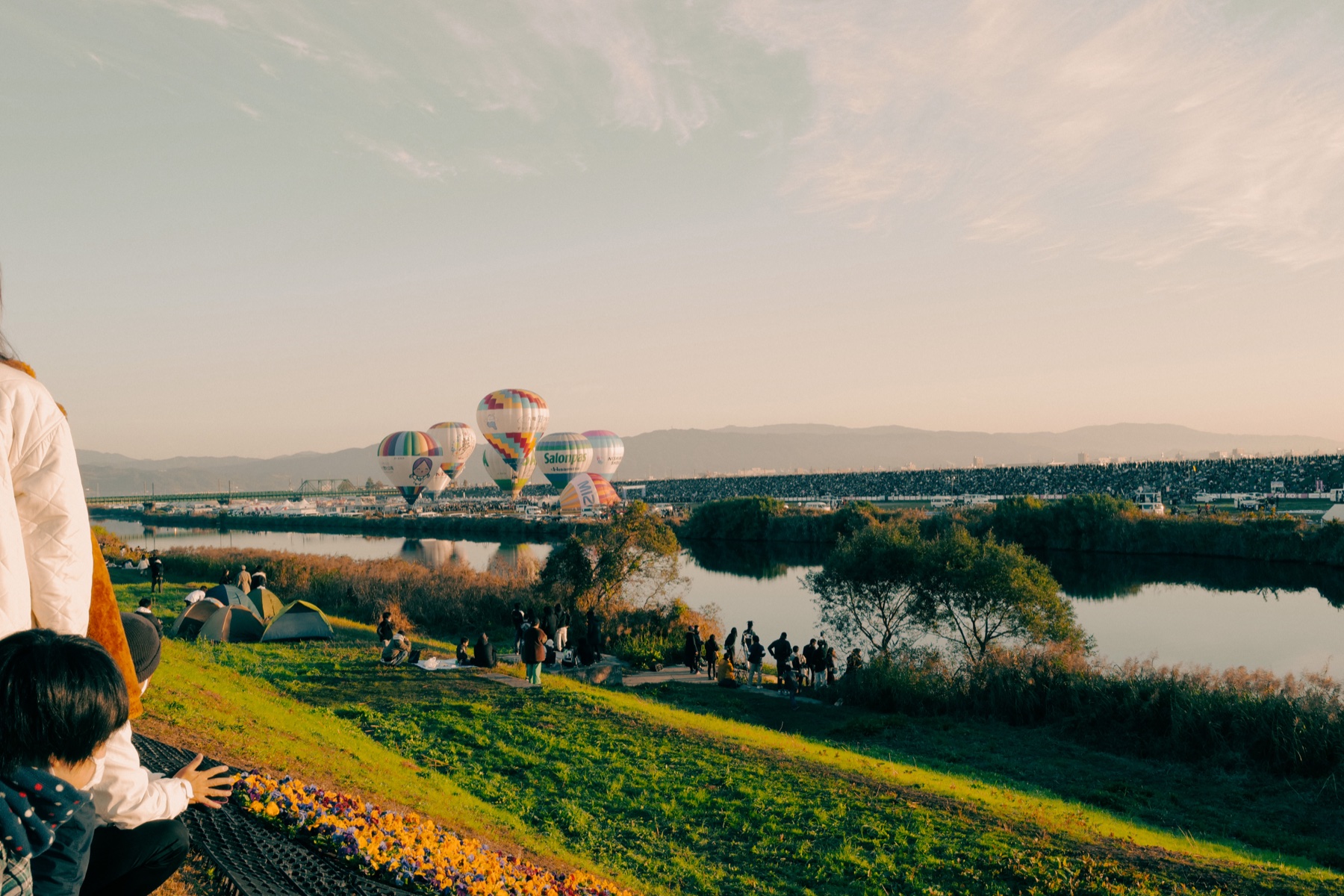 First balloons inflate in golden dawn light at the Saga International Balloon Fiesta, seen across the Kase River from the west bank — the dark mass of the main-venue crowd lines the far embankment while the near bank stays open.