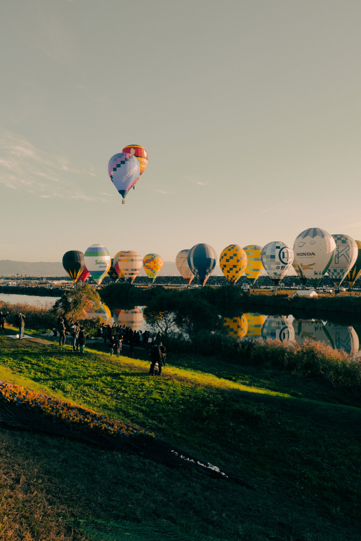 A balloon climbs into golden morning light above the launch rows at the Saga International Balloon Fiesta, with reflections in the Kase River and a handful of people watching from the grassy west bank slope in the foreground.