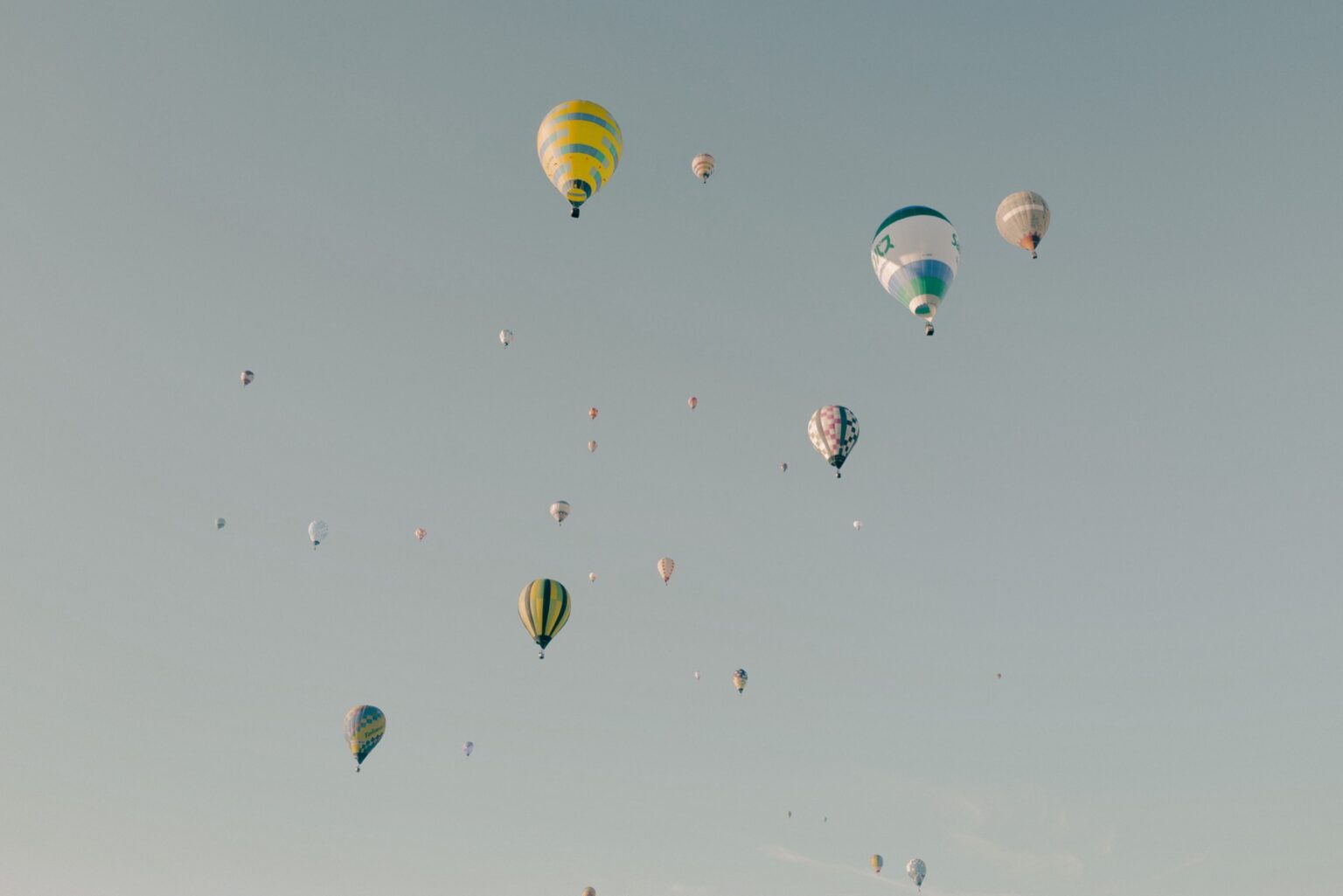 Dozens of hot air balloons scatter across a pale morning sky over the Saga plain after the mass ascent at the Saga International Balloon Fiesta, ranging from large nearby envelopes to tiny distant dots.