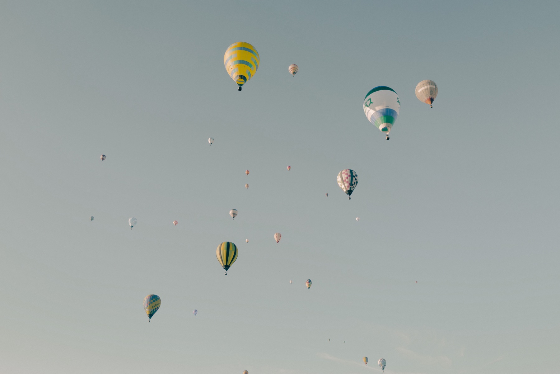 Dozens of hot air balloons scatter across a pale morning sky over the Saga plain after the mass ascent at the Saga International Balloon Fiesta, ranging from large nearby envelopes to tiny distant dots.