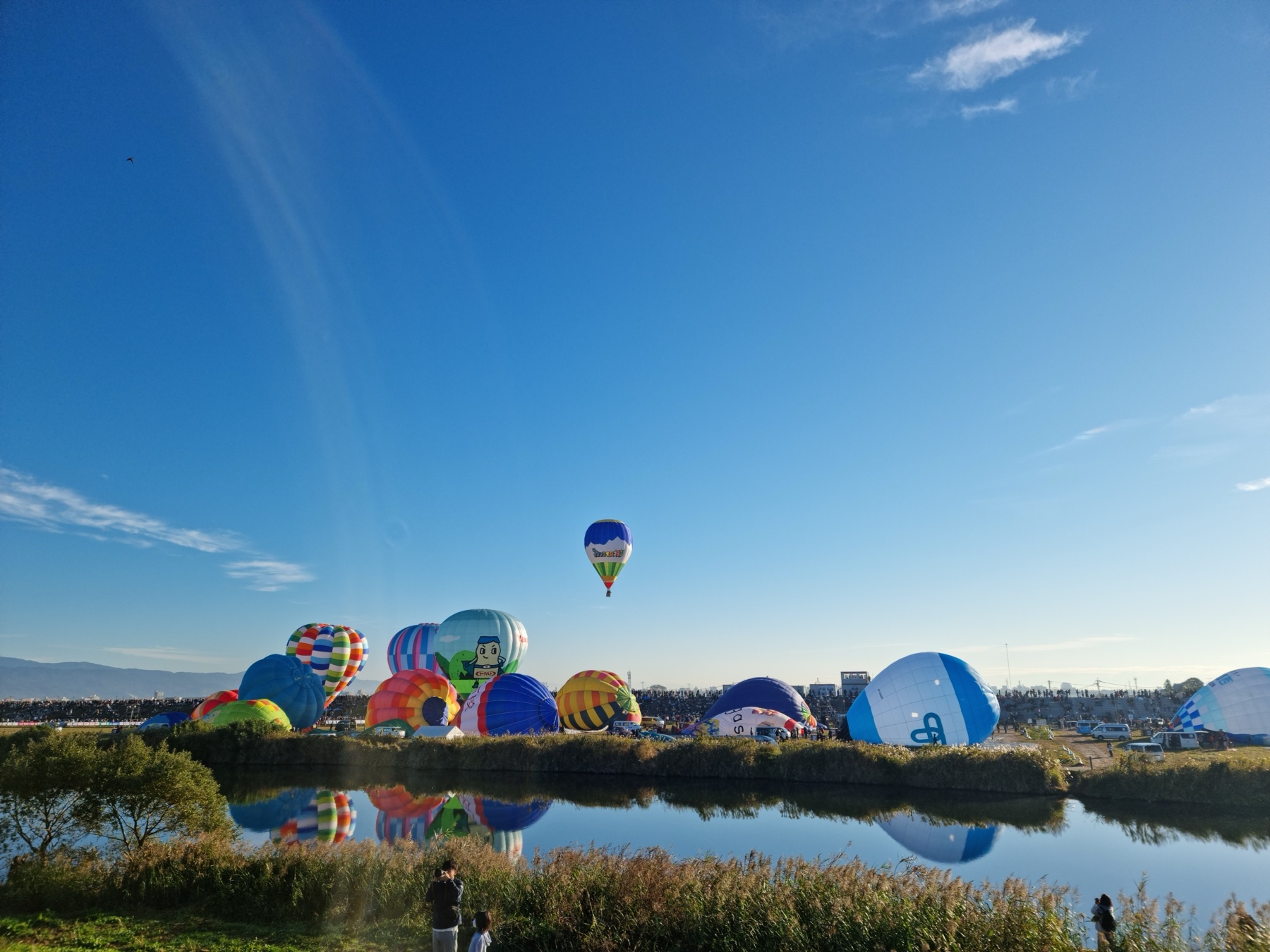 Cartoon-character and special-shape balloons inflate along the far bank of the Kase River under a deep blue sky at the Saga International Balloon Fiesta, with the whole colourful line-up reflected in the still water.