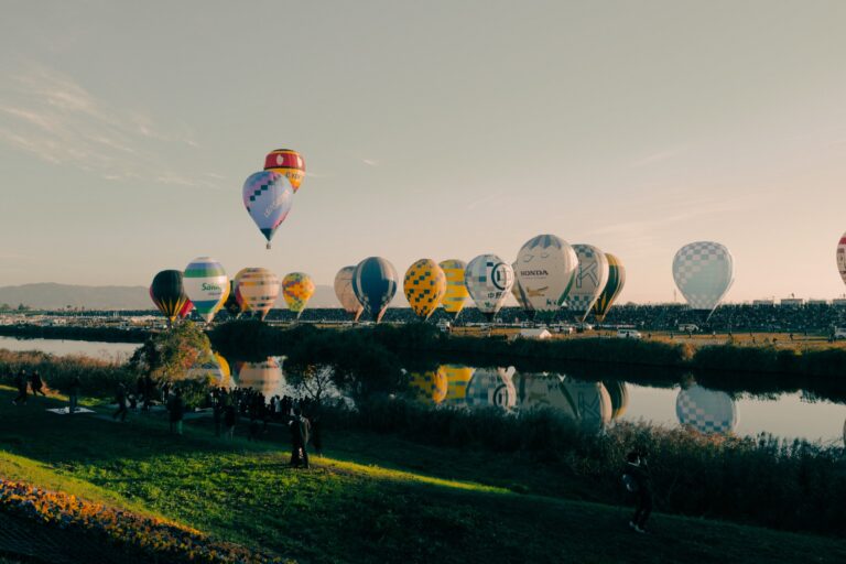 A hot air balloon lifts off above a long row of inflating balloons at the Saga International Balloon Fiesta, every envelope mirrored in the still morning water of the Kase River, photographed from the quiet west bank at sunrise.