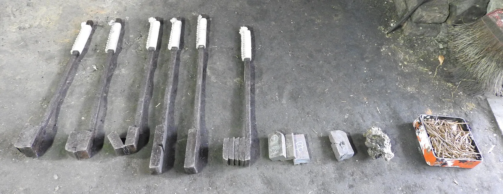 A swordsmith's forging tongs and billets of tatara steel and tamahagane laid out on a forge floor, the raw stages of making a Japanese blade