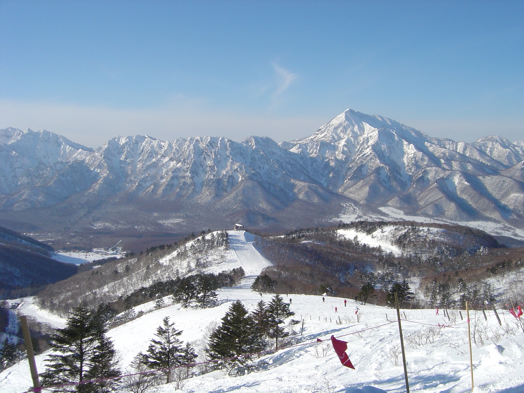 The jagged snow-covered ridgeline of the Togakushi mountains rises beyond a broad open snowfield and scattered pines at the Togakushi ski area near Nagano City.