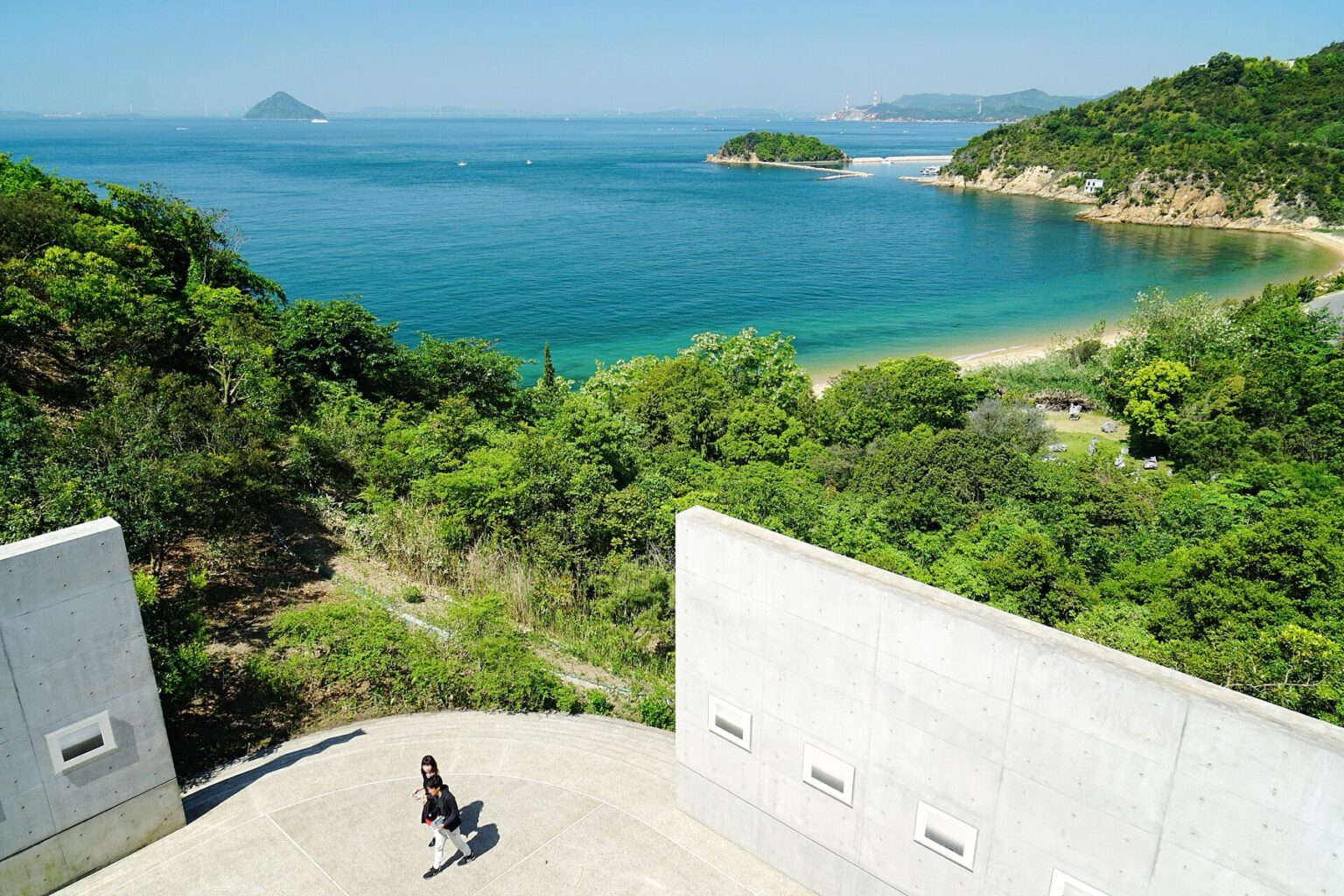 Two people walk across a circular raw concrete terrace at Benesse House Museum on Naoshima, framed by Tadao Ando's board-marked concrete walls with small square windows, looking out over wooded slopes to a turquoise bay and the islands of the Seto Inland Sea.