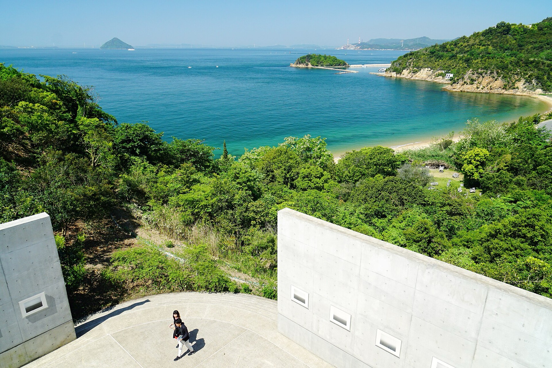 Two people walk across a circular raw concrete terrace at Benesse House Museum on Naoshima, framed by Tadao Ando's board-marked concrete walls with small square windows, looking out over wooded slopes to a turquoise bay and the islands of the Seto Inland Sea.