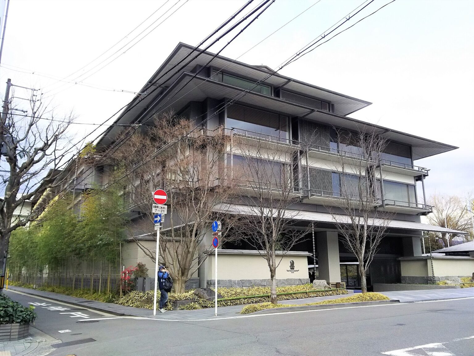 The low-rise exterior of The Ritz-Carlton Kyoto, showing deep overhanging eaves in dark timber, balconies screened with vertical louvres, a bamboo hedge along the pavement and the hotel's lion crest on a low stone-faced wall.
