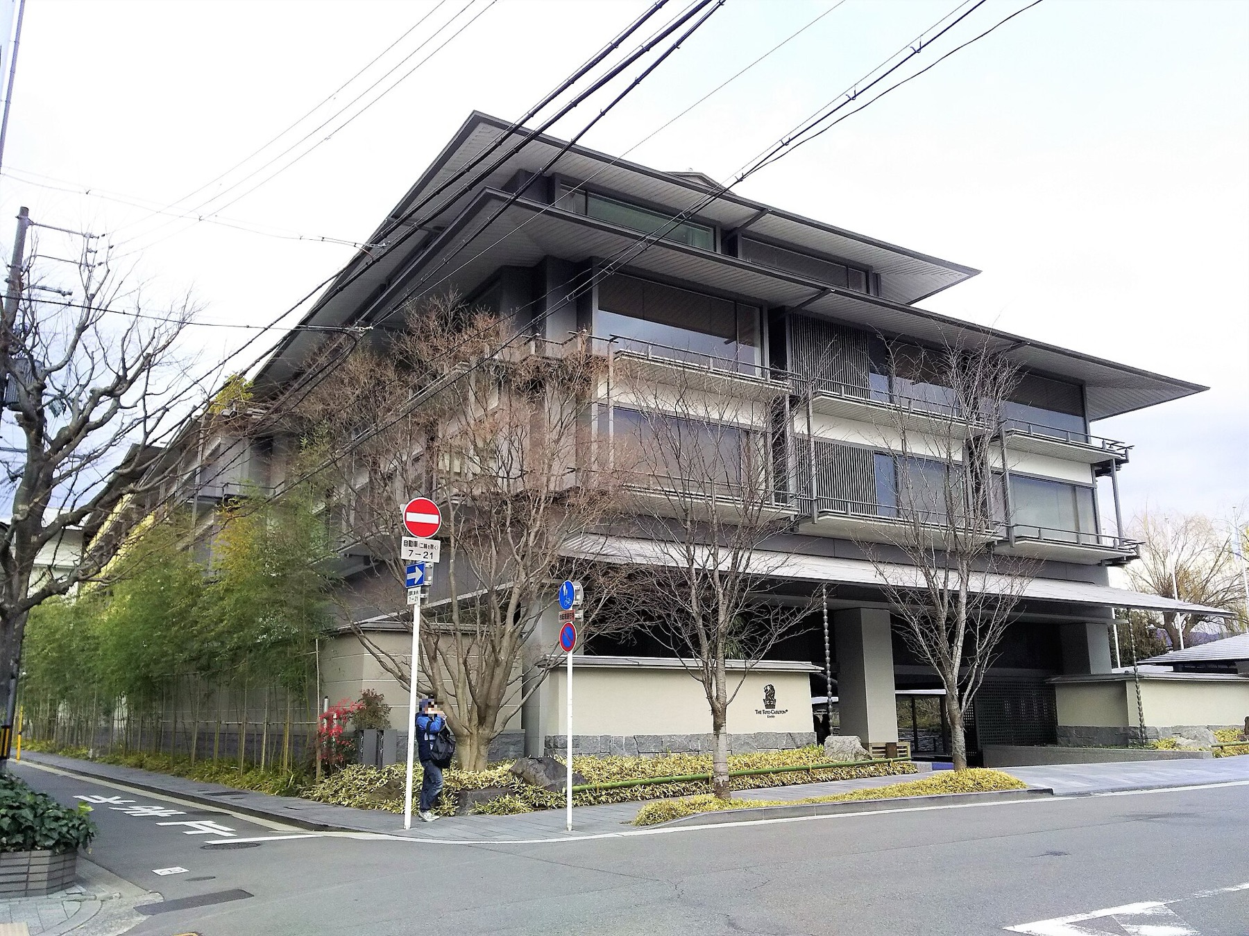 The low-rise exterior of The Ritz-Carlton Kyoto, showing deep overhanging eaves in dark timber, balconies screened with vertical louvres, a bamboo hedge along the pavement and the hotel's lion crest on a low stone-faced wall.