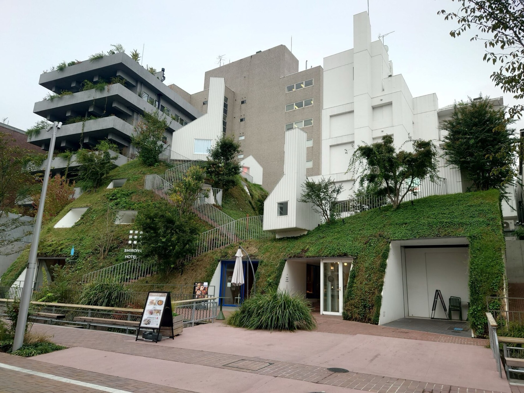 The grass-covered artificial hill of Shiroiya Hotel's Green Tower in Maebashi, Gunma, with white cabin-like guest rooms embedded in the slope, planted terraces stepping up the concrete Heritage Tower on the left and a staircase climbing the hill.