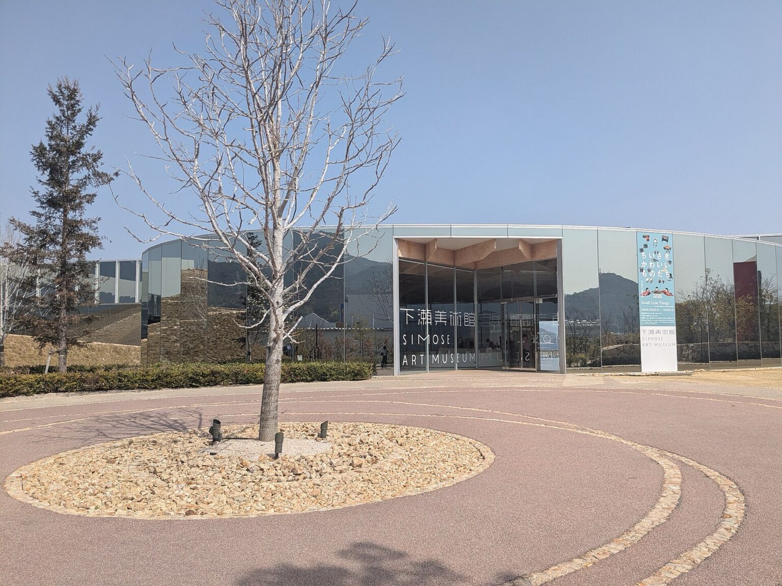 The mirrored glass entrance pavilion of the Simose Art Museum reflects the surrounding hills and a stone wall, with bilingual signage reading Simose Art Museum in Japanese and English and a bare tree in a circular gravel bed in the forecourt.