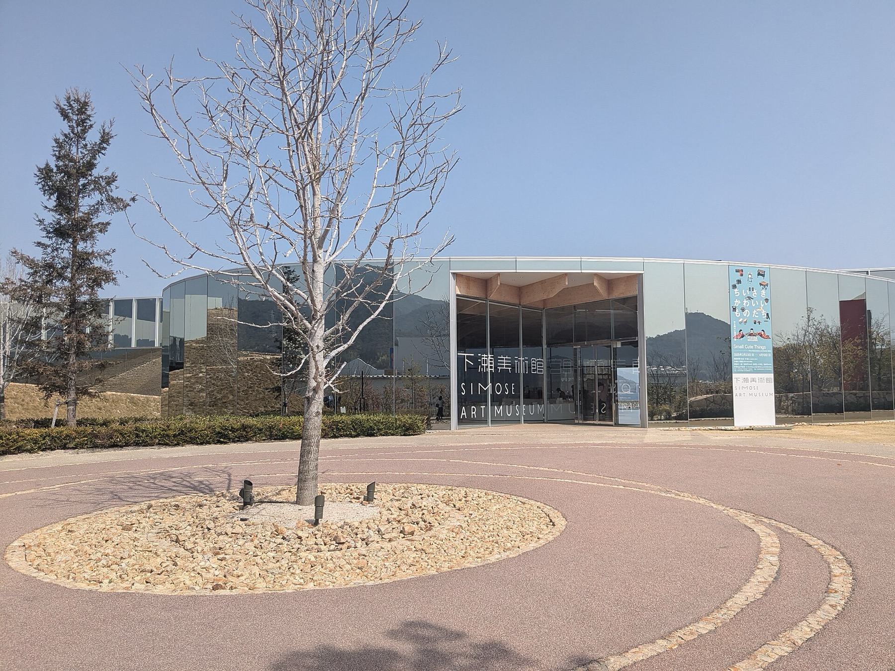 The mirrored glass entrance pavilion of the Simose Art Museum reflects the surrounding hills and a stone wall, with bilingual signage reading Simose Art Museum in Japanese and English and a bare tree in a circular gravel bed in the forecourt.