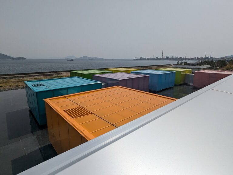 Eight brightly coloured movable gallery boxes in orange, teal, purple, green, yellow and pink float on a shallow reflecting water basin at the Simose Art Museum in Otake, Hiroshima, with the Seto Inland Sea, distant islands and the cranes of an industrial waterfront behind them under a hazy sky.
